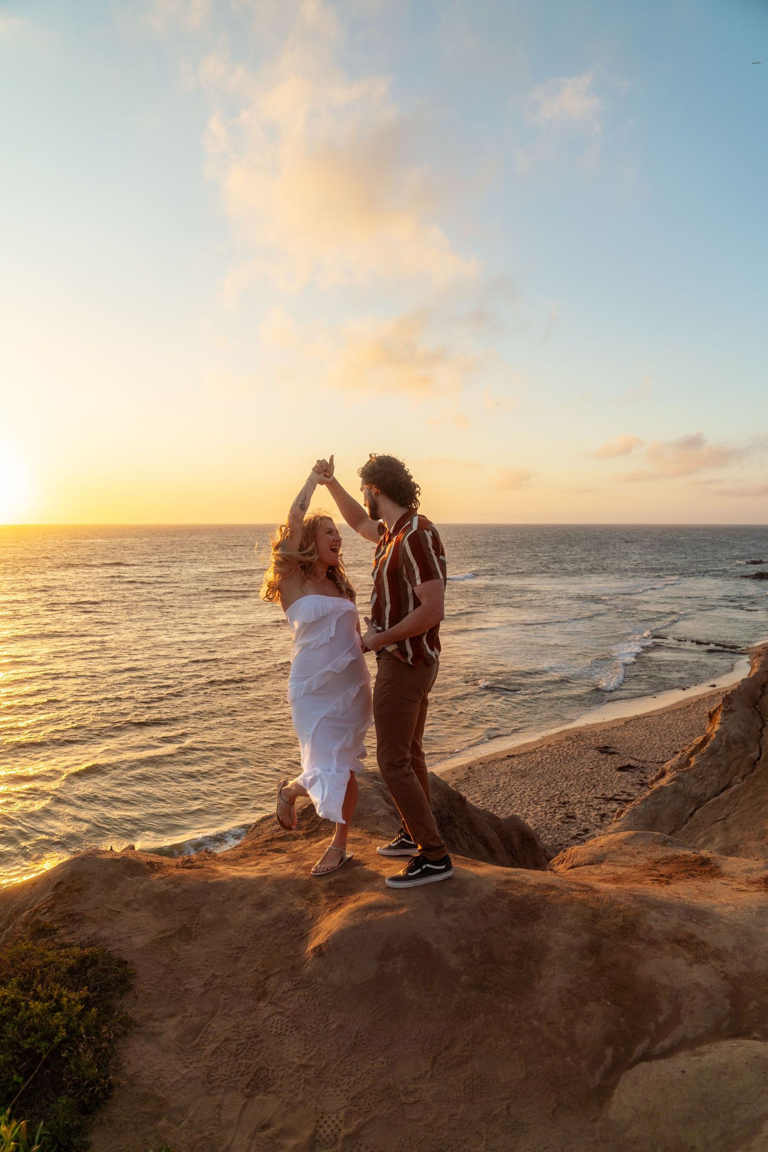 Couple dancing together on coastal cliff overlooking ocean at sunset with golden sky.