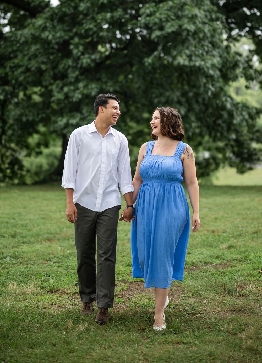 A couple walking and holding hands in a park with green trees, the woman wearing a blue summer dress.
