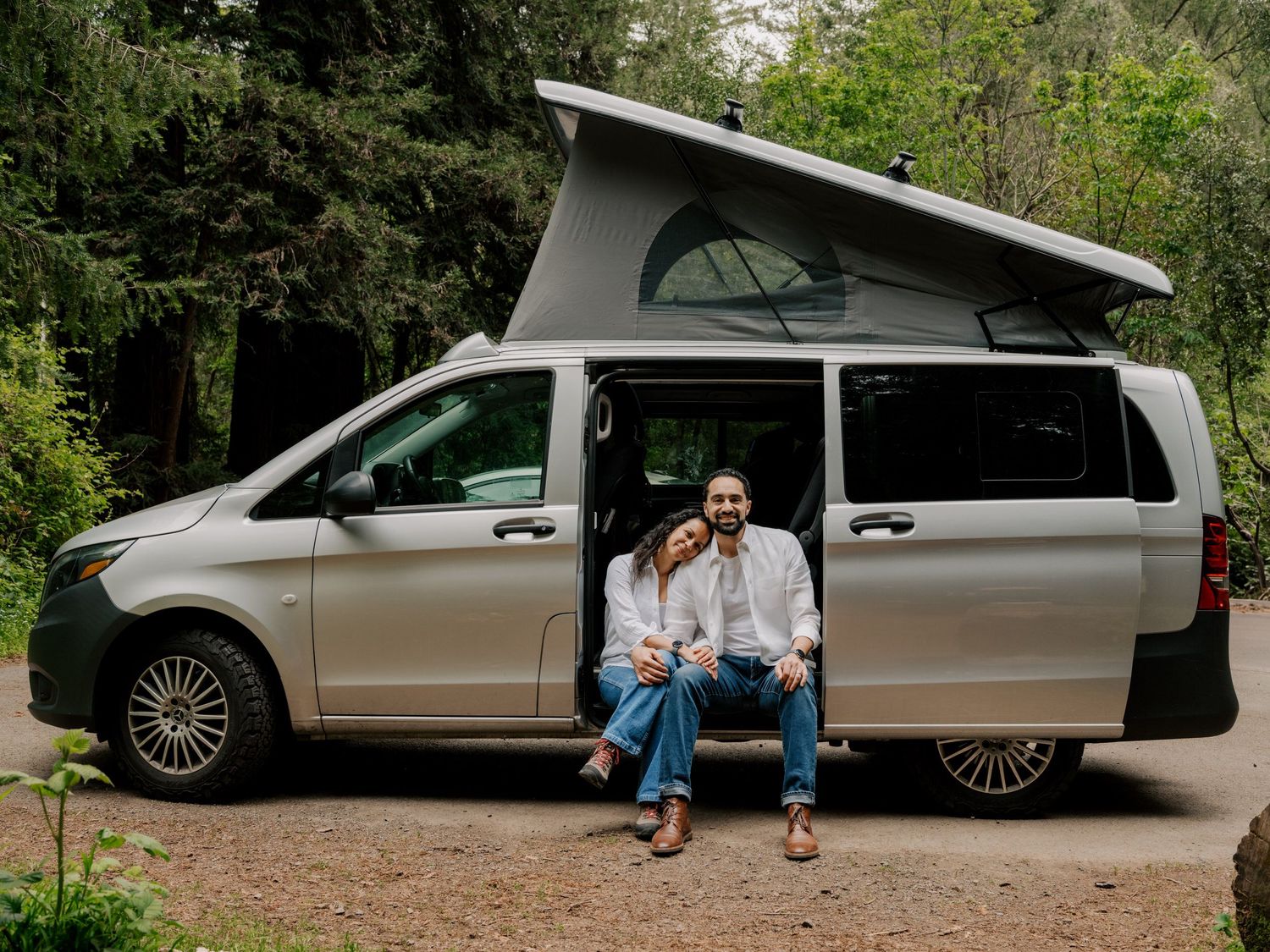 A silver Mercedes Metris camper van with pop-up roof parked in a wooded area while its owners sit in the doorway.
