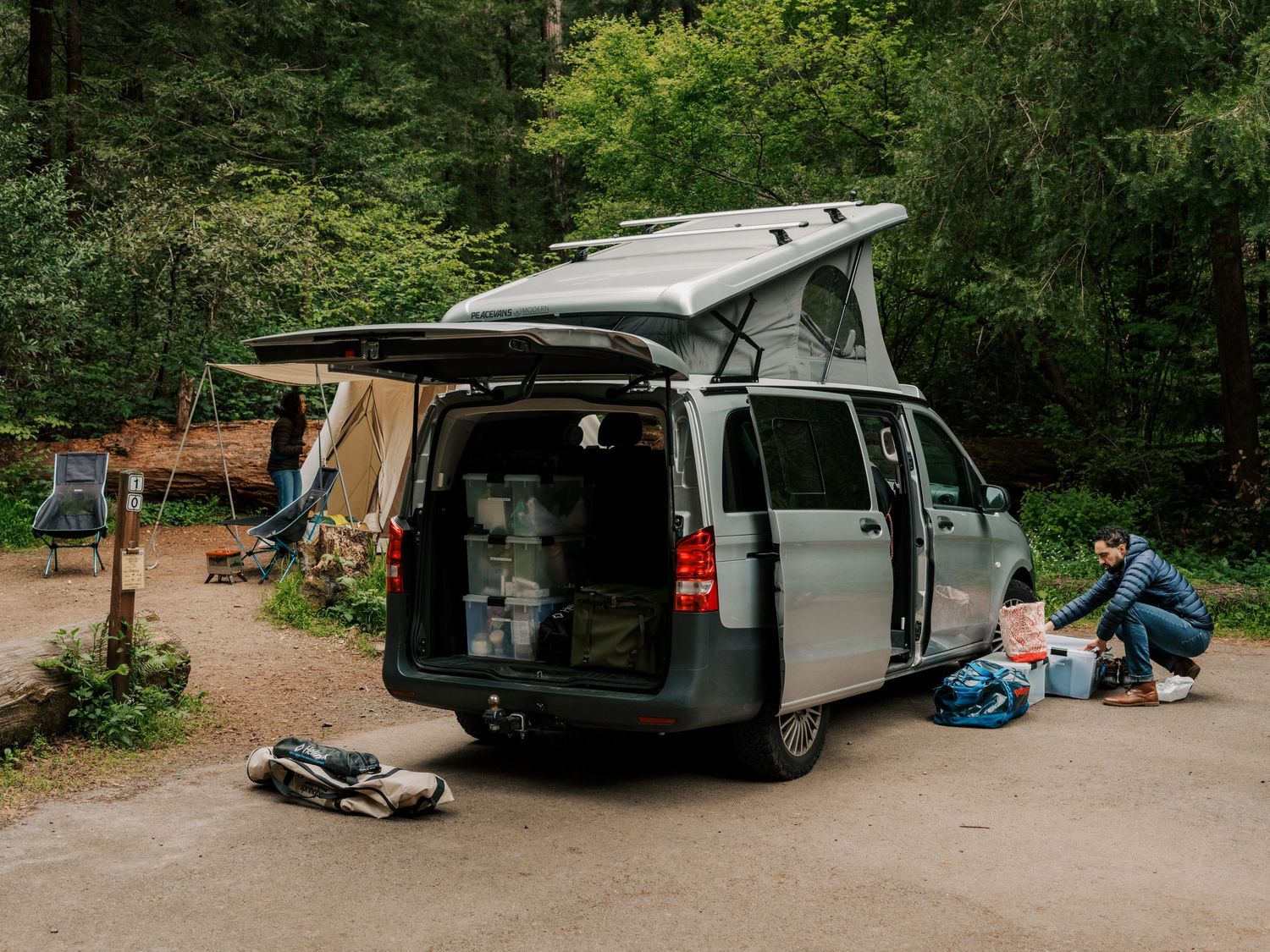 A camper van with pop-up roof and opened side door parked at a wooded campsite with camping gear spread around it.