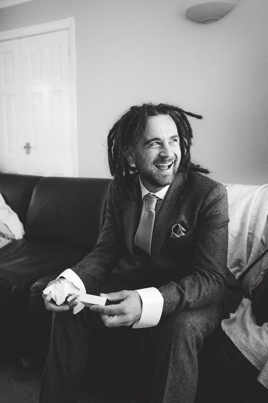A person wearing a suit and tie laughs while sitting on a couch holding a game controller in a black and white photo.