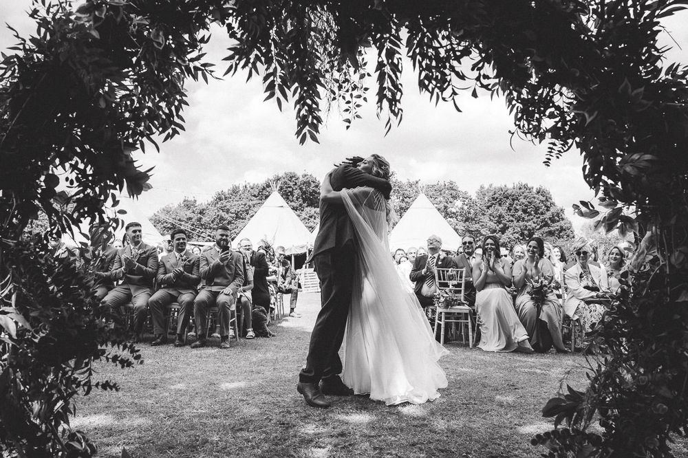 Black and white photo of couple dancing at outdoor garden wedding reception surrounded by guests under tent and trees.