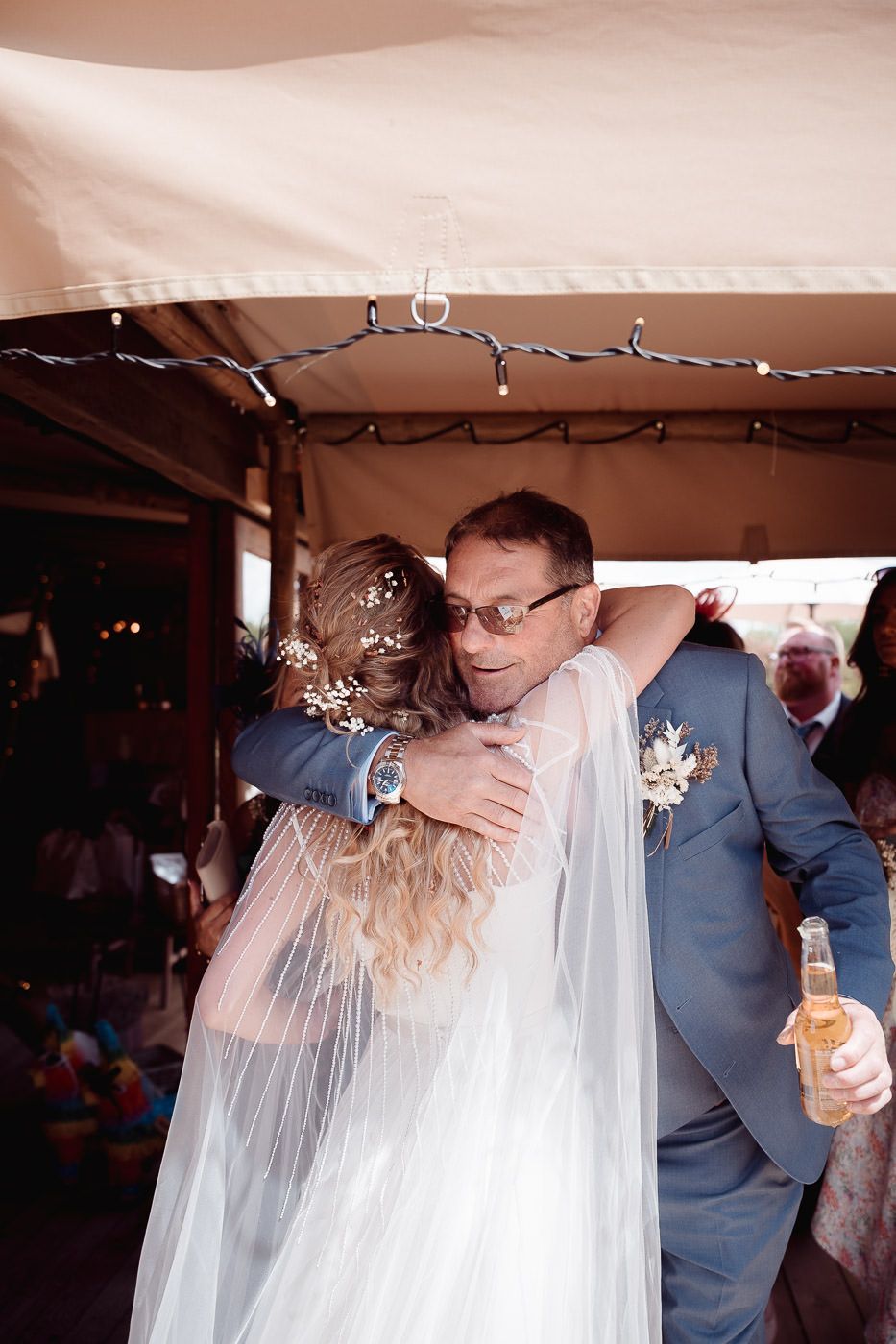 A tender embrace between a bride in flowing veil and person in gray suit during an outdoor wedding celebration.