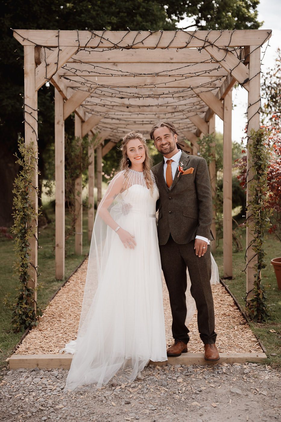 Newlywed couple stands in romantic garden pergola decorated with draped fabric and twinkle lights on wedding day.