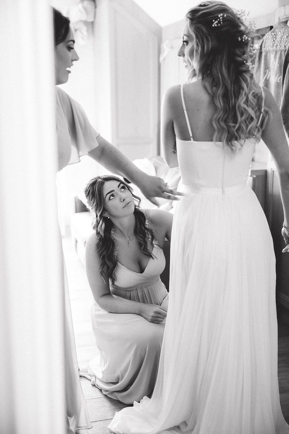 Black and white photo of bridesmaids helping adjust a bride's flowing white wedding gown during preparation.
