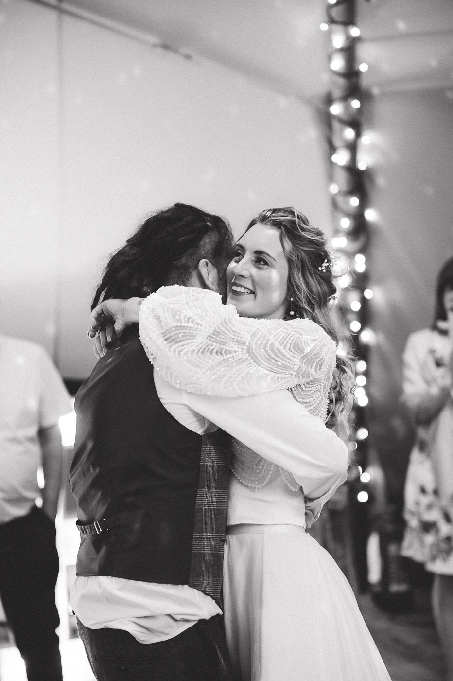 A black and white candid photo of a romantic dance embrace at a wedding reception with string lights in the background.