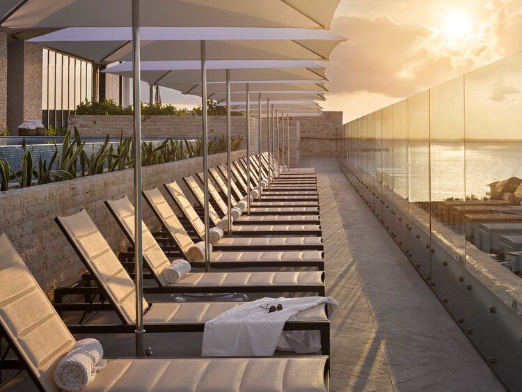 Row of elegant lounge chairs on luxury hotel rooftop terrace overlooking sunset through glass panels.