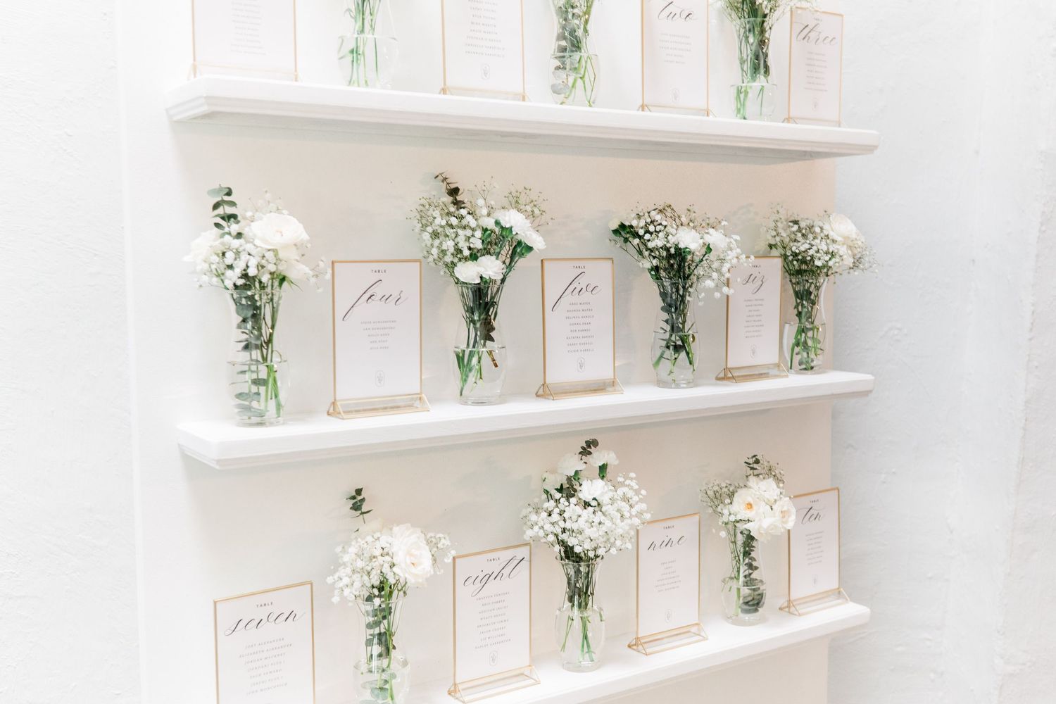 White floating shelves display elegant glass vases filled with delicate white flowers and greenery alongside pink-framed cards.