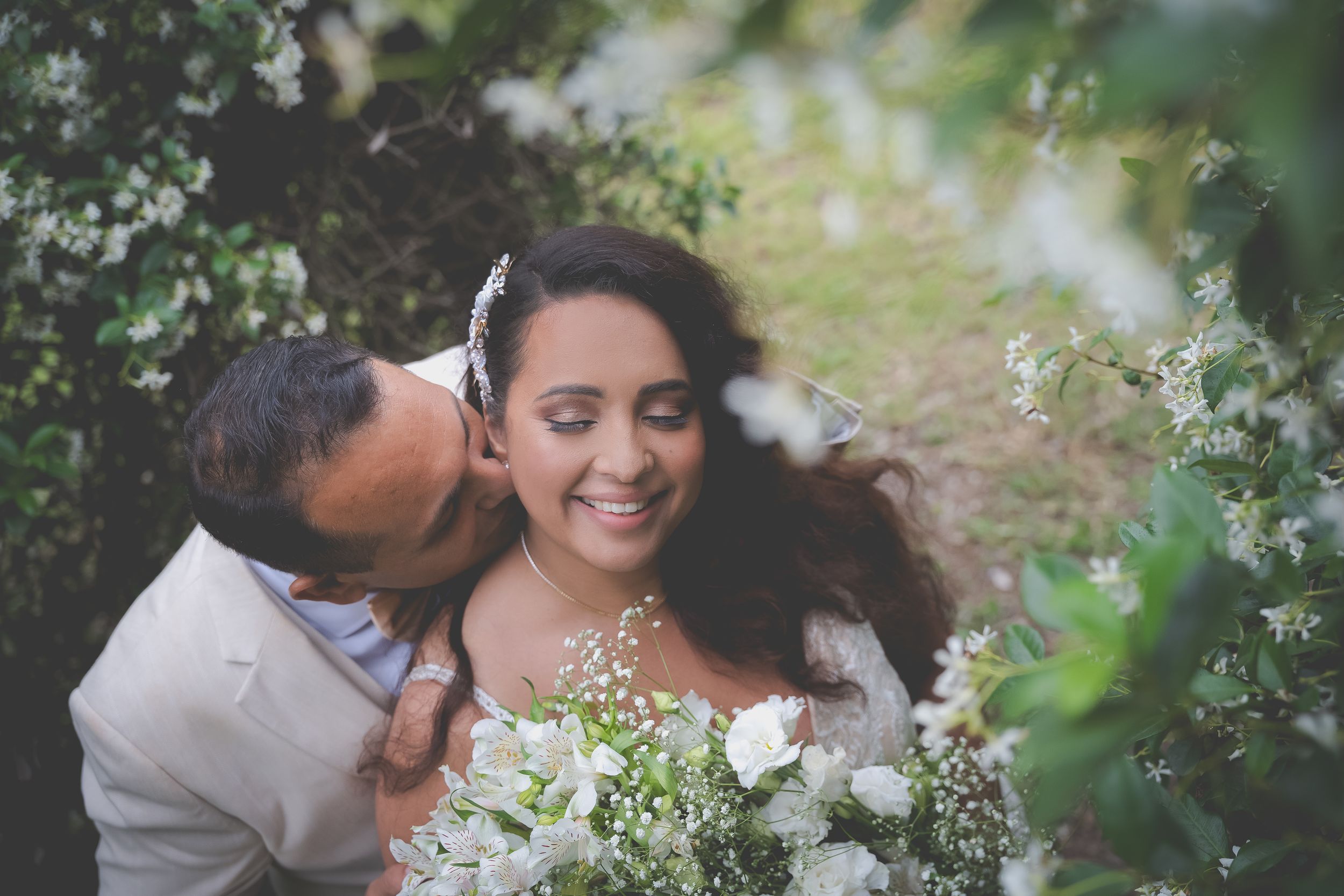 A wedding photo, the groom kissing the cheek of the bride with her eyes closed and surrounded by flowers