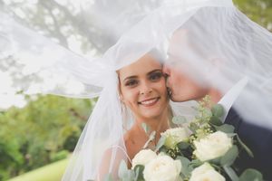 A groom kisses his bride who looks at the camera smiling both hidden under the brides veil.