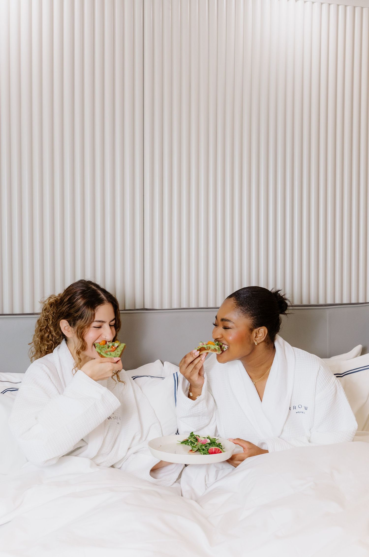 Two people in white bathrobes engage in a playful pillow fight on a hotel bed next to large windows.
