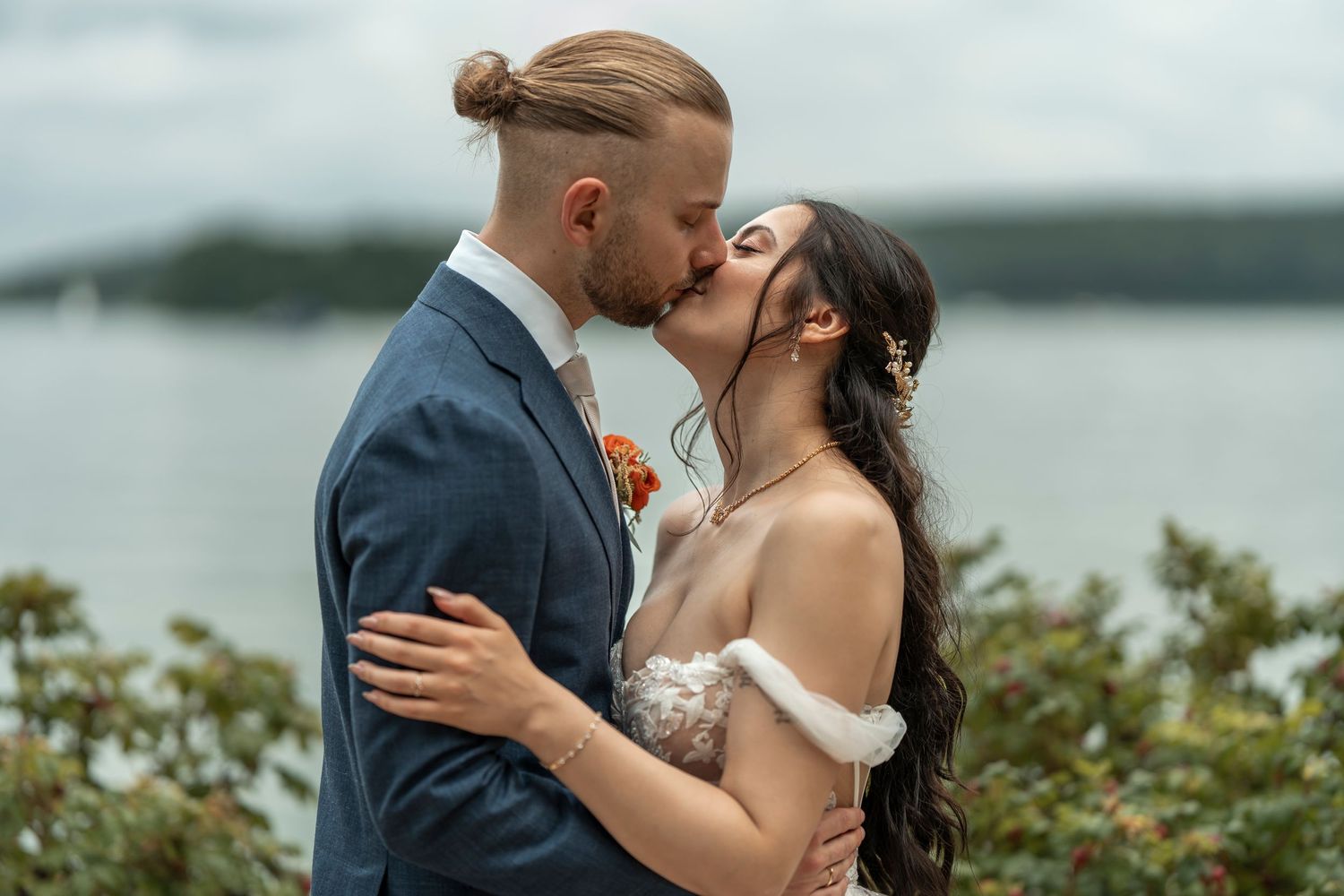 Ein Paar teilt einen romantischen Kuss an ihrem Hochzeitstag, mit einem See und Grün im Hintergrund.