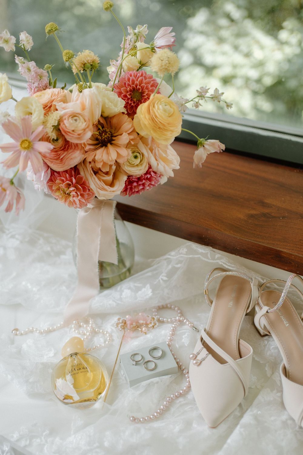 Elegant bridal flat lay featuring a pink and yellow bouquet, pearl necklace, wedding rings, perfume, and ivory heels on lace.