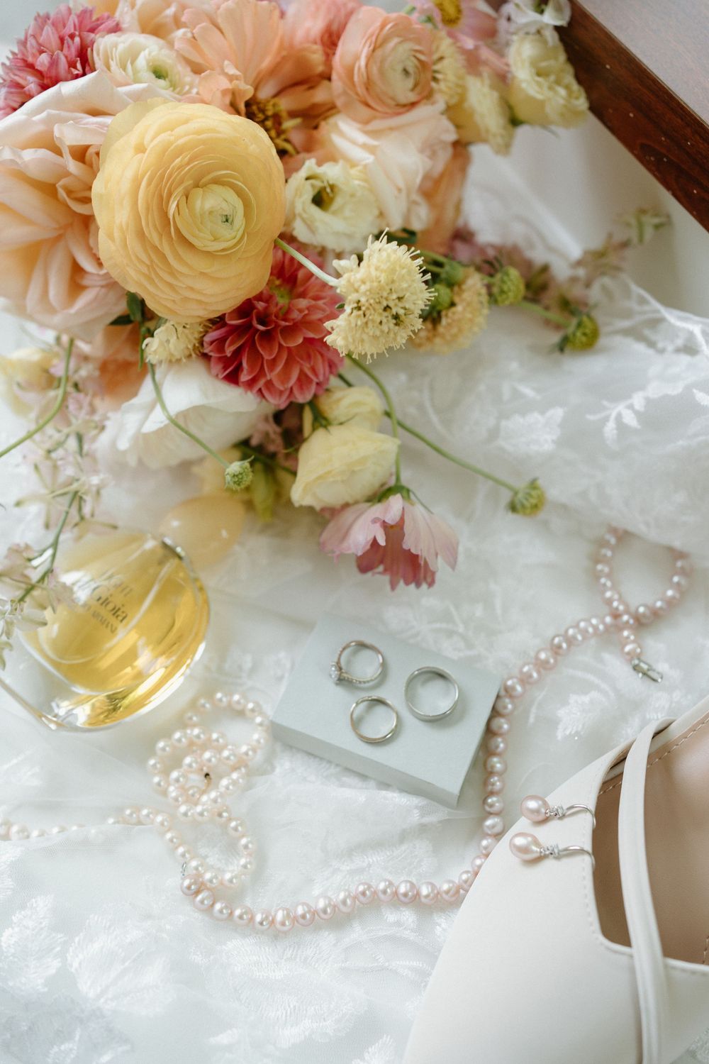 Close-up bridal detail shot with colorful ranunculus bouquet, pearl necklace, wedding bands, and perfume bottle on white lace.
