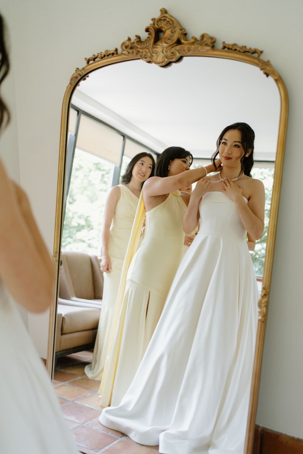Bride in strapless white gown getting necklace fastened by bridesmaid in yellow dress, reflected in ornate gold mirror.
