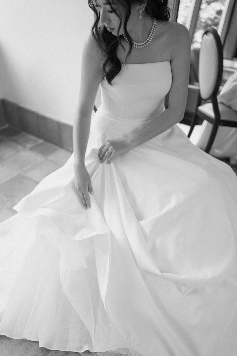 Black and white portrait of bride in strapless ball gown with pearl necklace, gracefully arranging her flowing skirt.