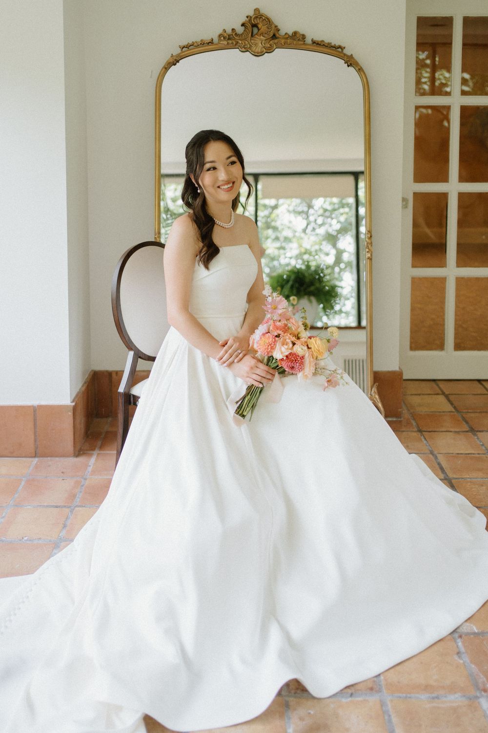 Smiling bride in strapless white satin ball gown holding a pink bouquet, posing before an ornate gold-framed mirror.
