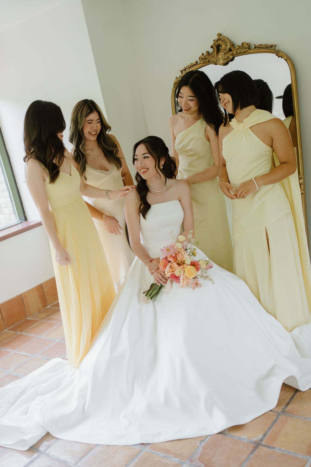 Bride in white strapless gown holding pink bouquet surrounded by laughing bridesmaids in soft yellow dresses near gold mirror.