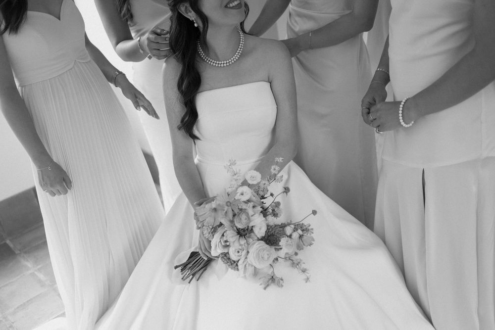 Black and white close-up of bride in pearl necklace holding a lush bouquet, surrounded by bridesmaids in flowing gowns.