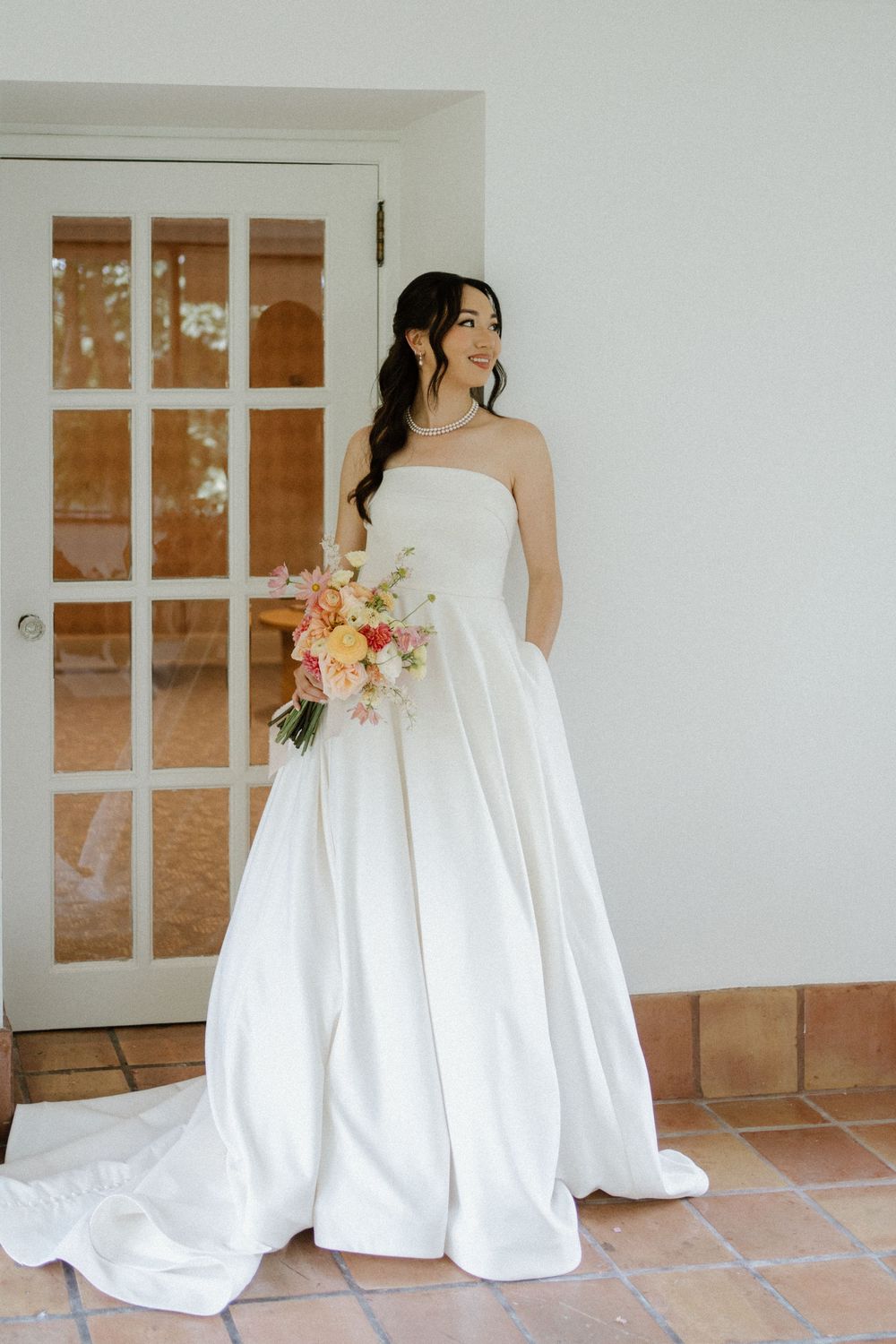 Elegant bride in strapless white satin ball gown with long train holding a colorful pink and yellow bouquet indoors.