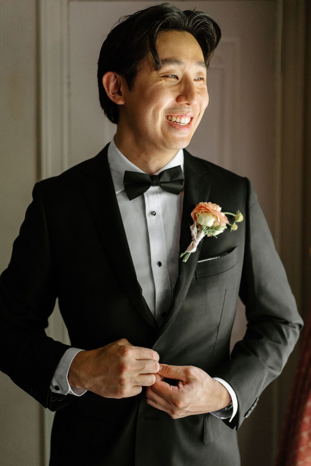 Smiling groom in black tuxedo adjusts jacket button wearing floral boutonniere on wedding day.