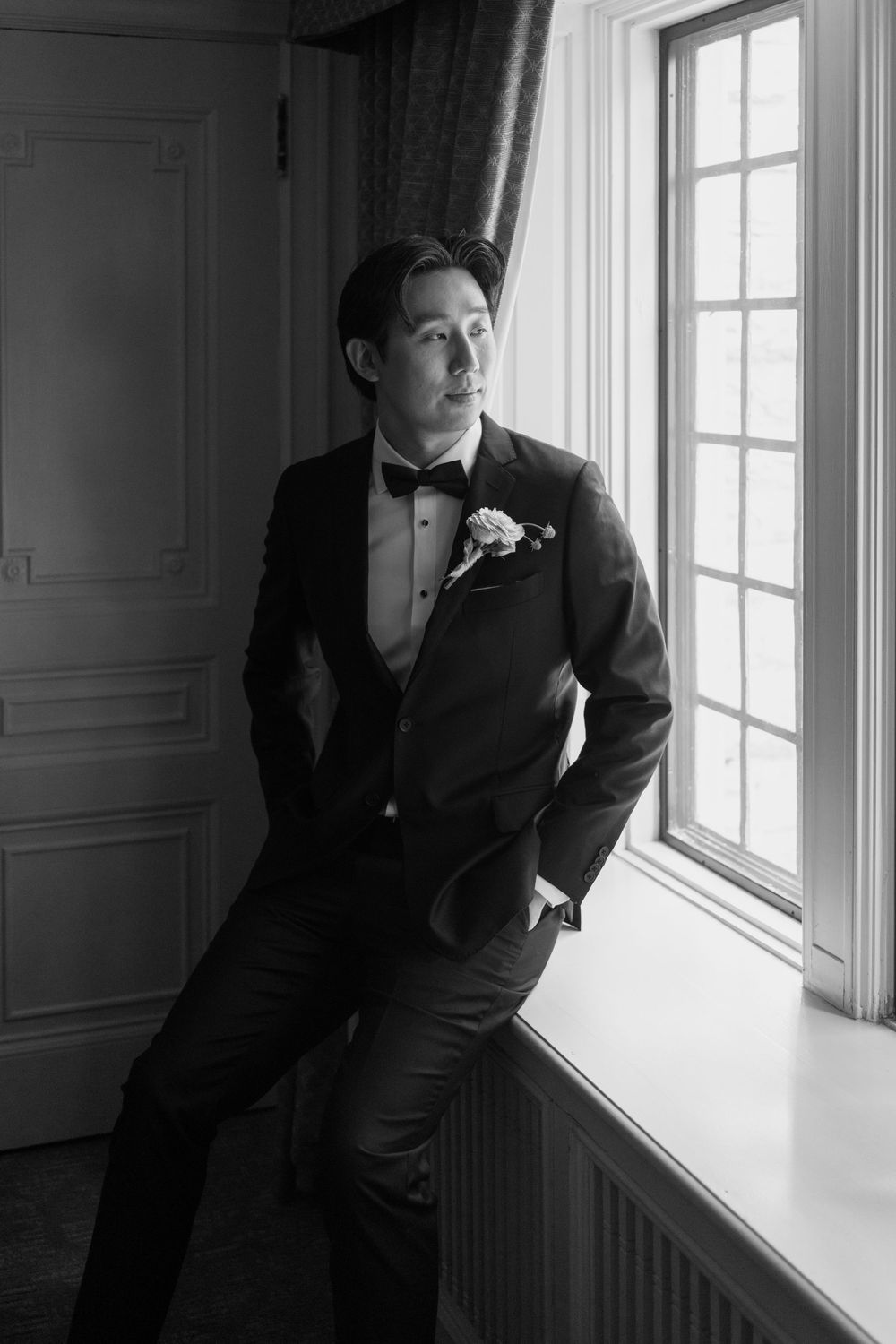 Dapper groom in black tuxedo with boutonniere poses by window in classic black and white wedding portrait.