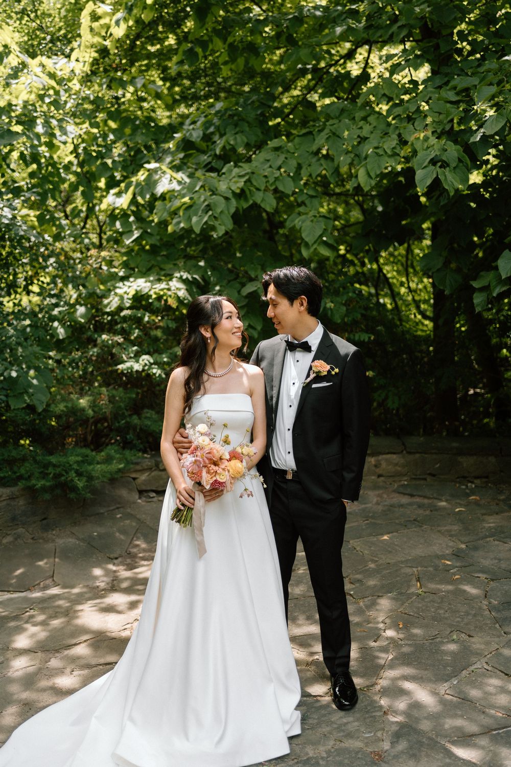 Bride and groom share a joyful moment outdoors holding a colorful bouquet surrounded by lush green trees.