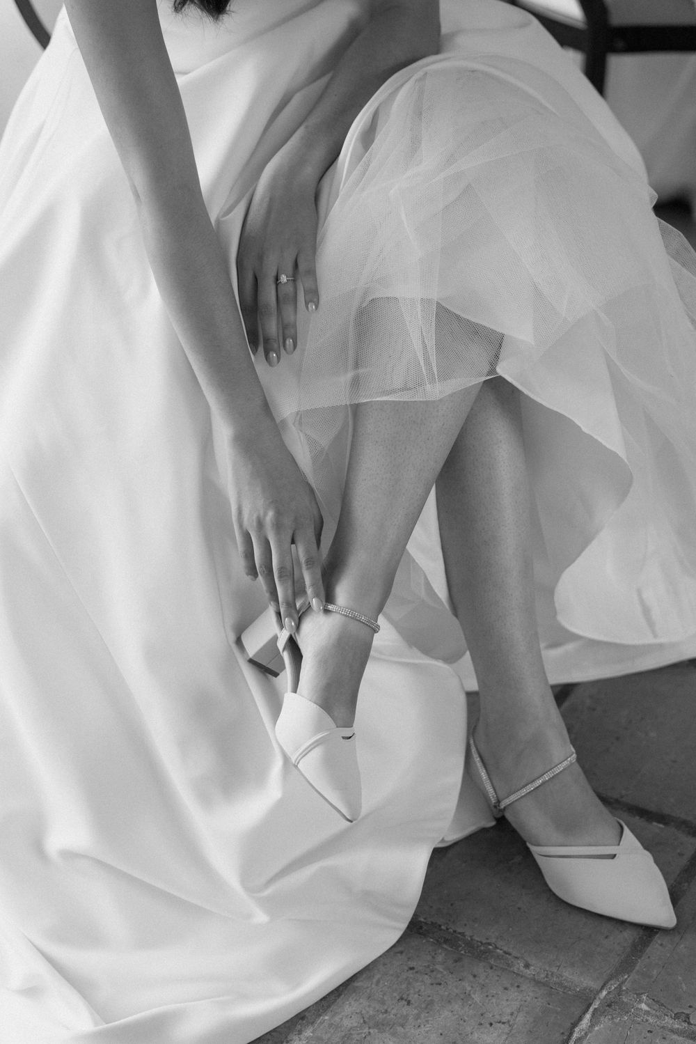Black and white close-up of a bride adjusting her white pointed-toe heels beneath her wedding gown.