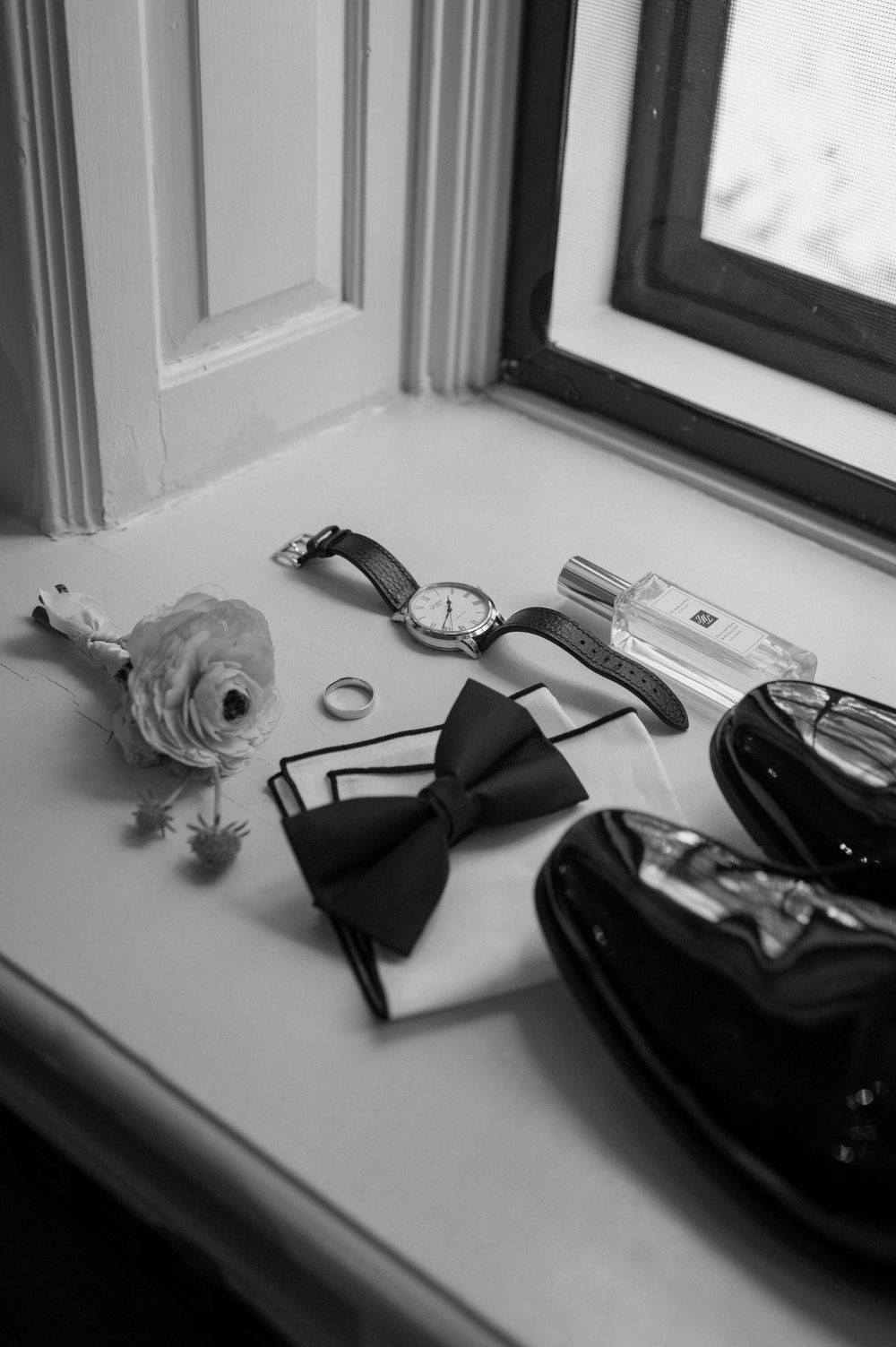 Black and white flat lay of groom wedding accessories including bow tie, watch, ring, boutonniere, and cologne on windowsill.