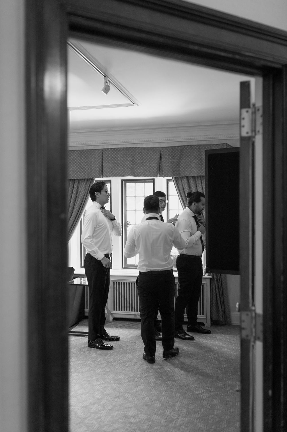Black and white photo of groomsmen getting ready in elegant room viewed through doorway.