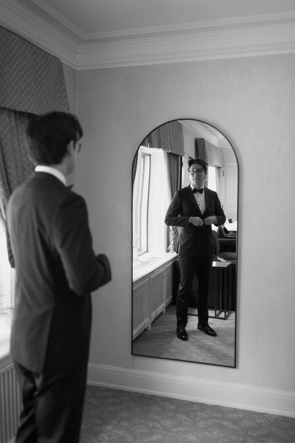 Black and white photo of groom in tuxedo checking appearance in arched full-length mirror.