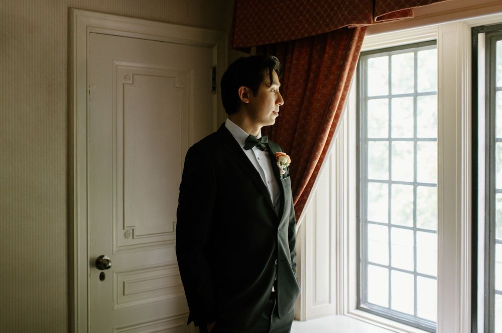 Groom in black tuxedo with bow tie gazing out window in elegant room before wedding ceremony.