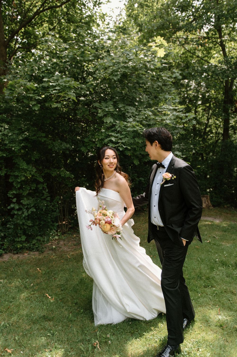 Bride in strapless white gown holding colorful bouquet smiles at groom in black tuxedo outdoors.