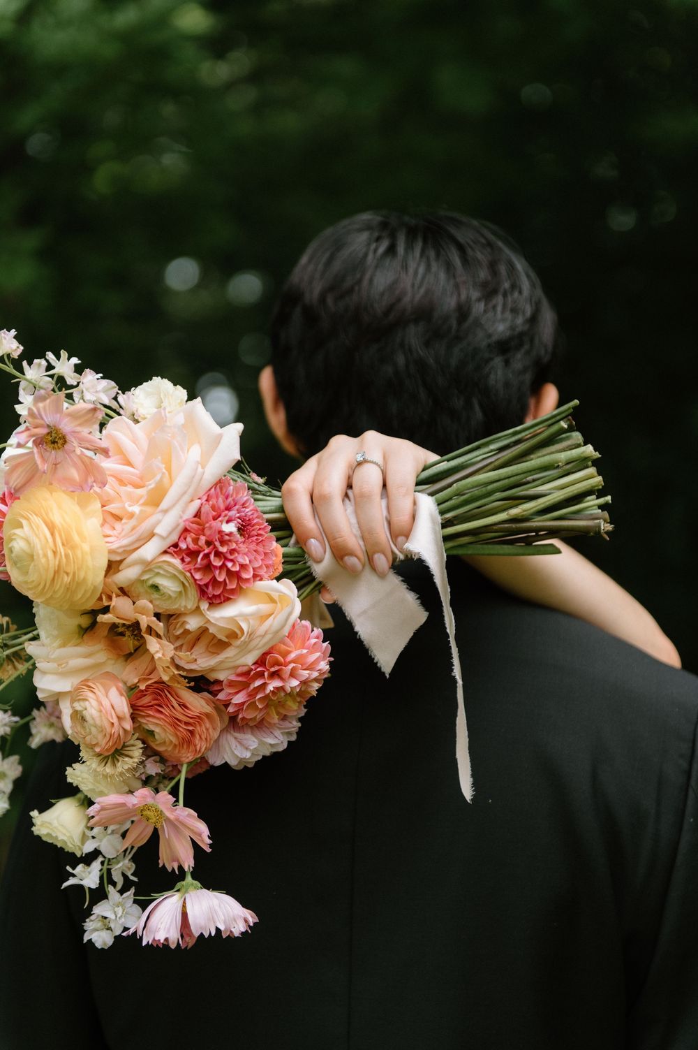 Close-up of bride holding lush pink and coral wedding bouquet over groom's shoulder in garden.