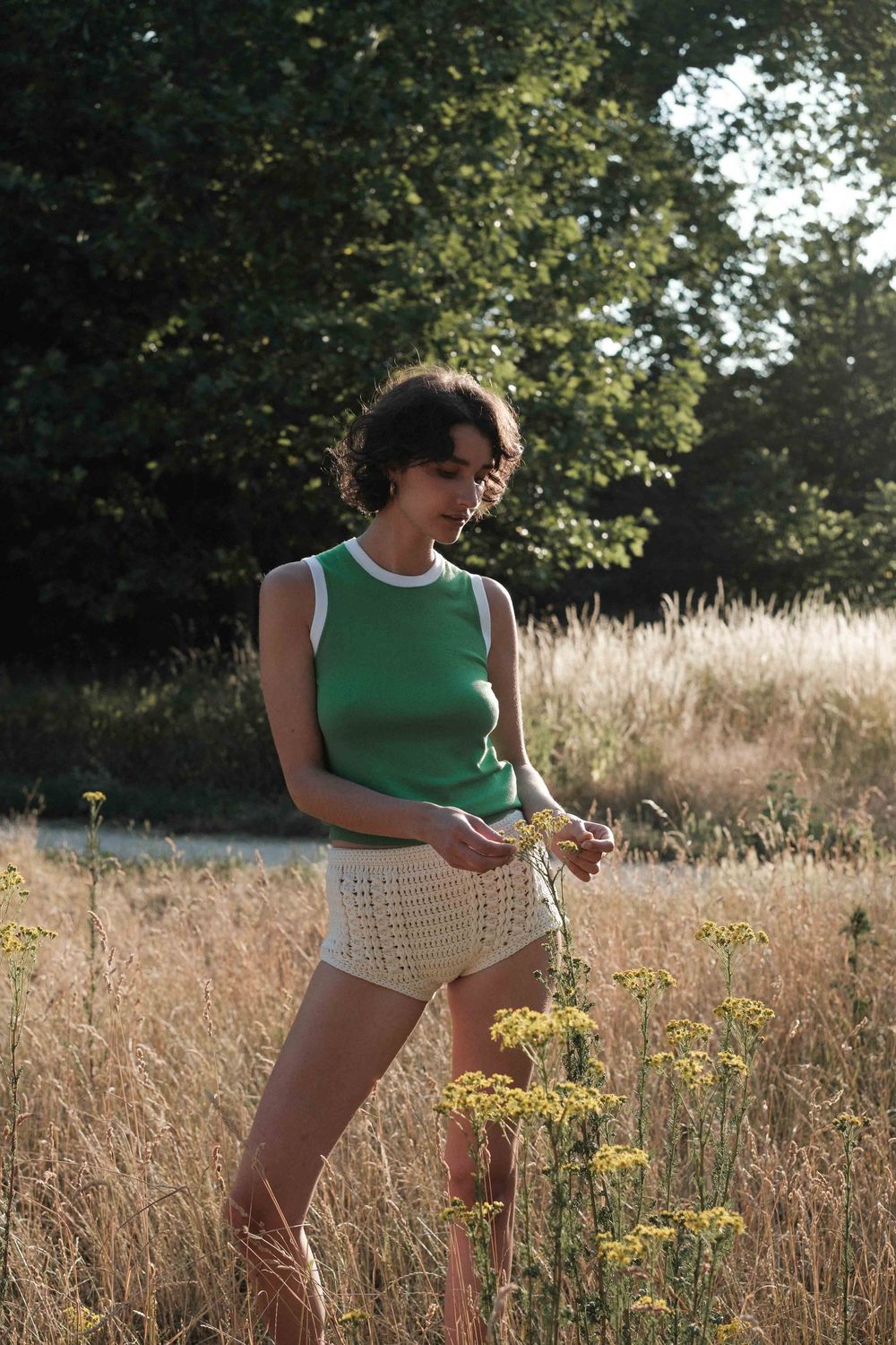 A person wearing a green tank top and white knit top stands in a golden field of tall grasses on a sunny day.