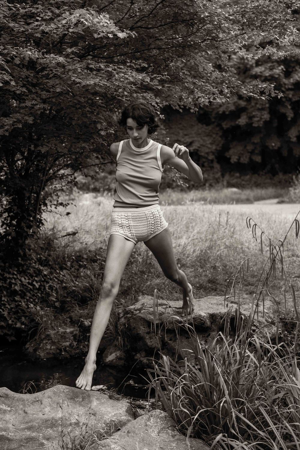Black and white nature photo showing someone in summer attire jumping on rocks by a stream in a wooded area.