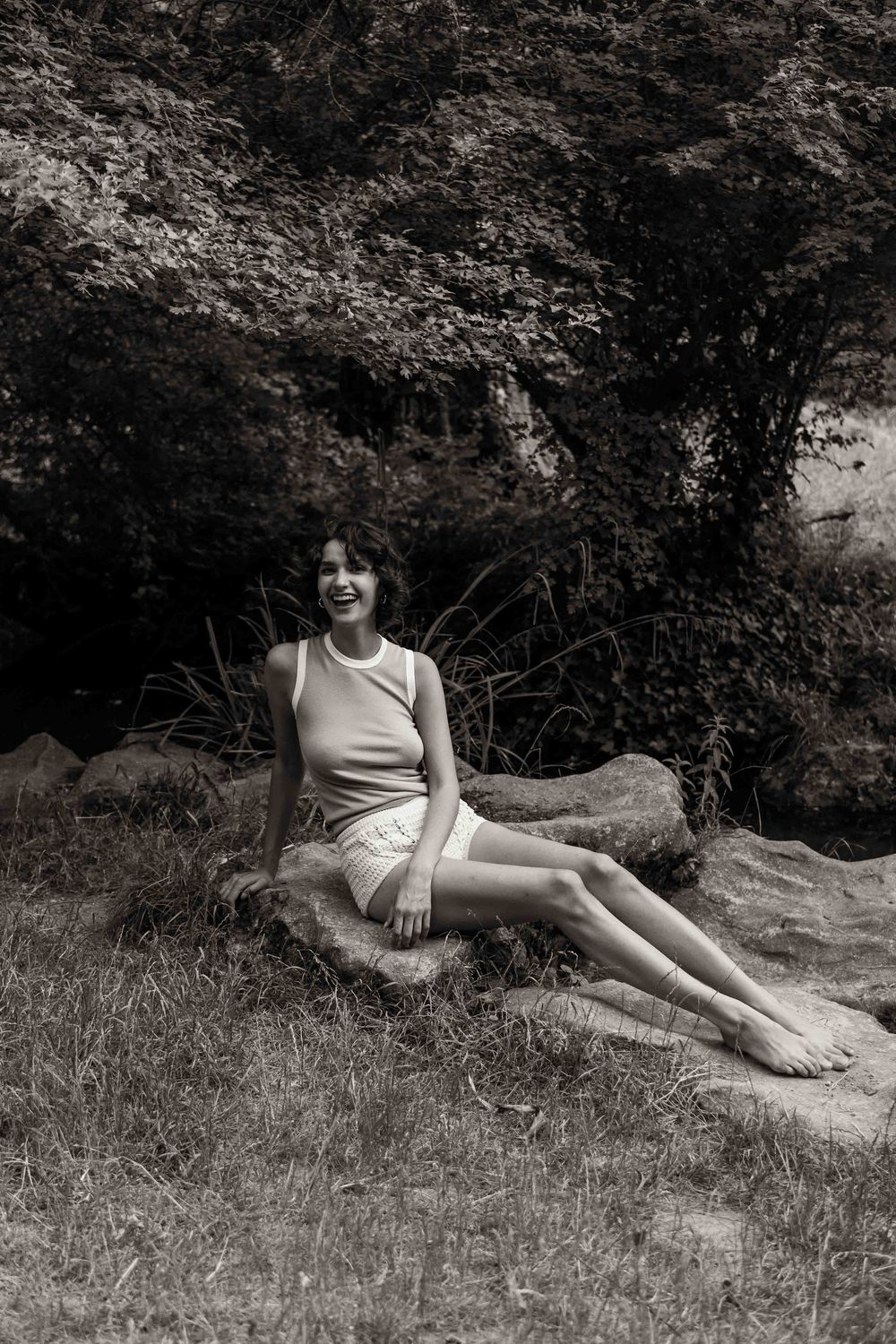 Black and white vintage-style photo showing someone relaxing on a rock in summer attire with trees in background.