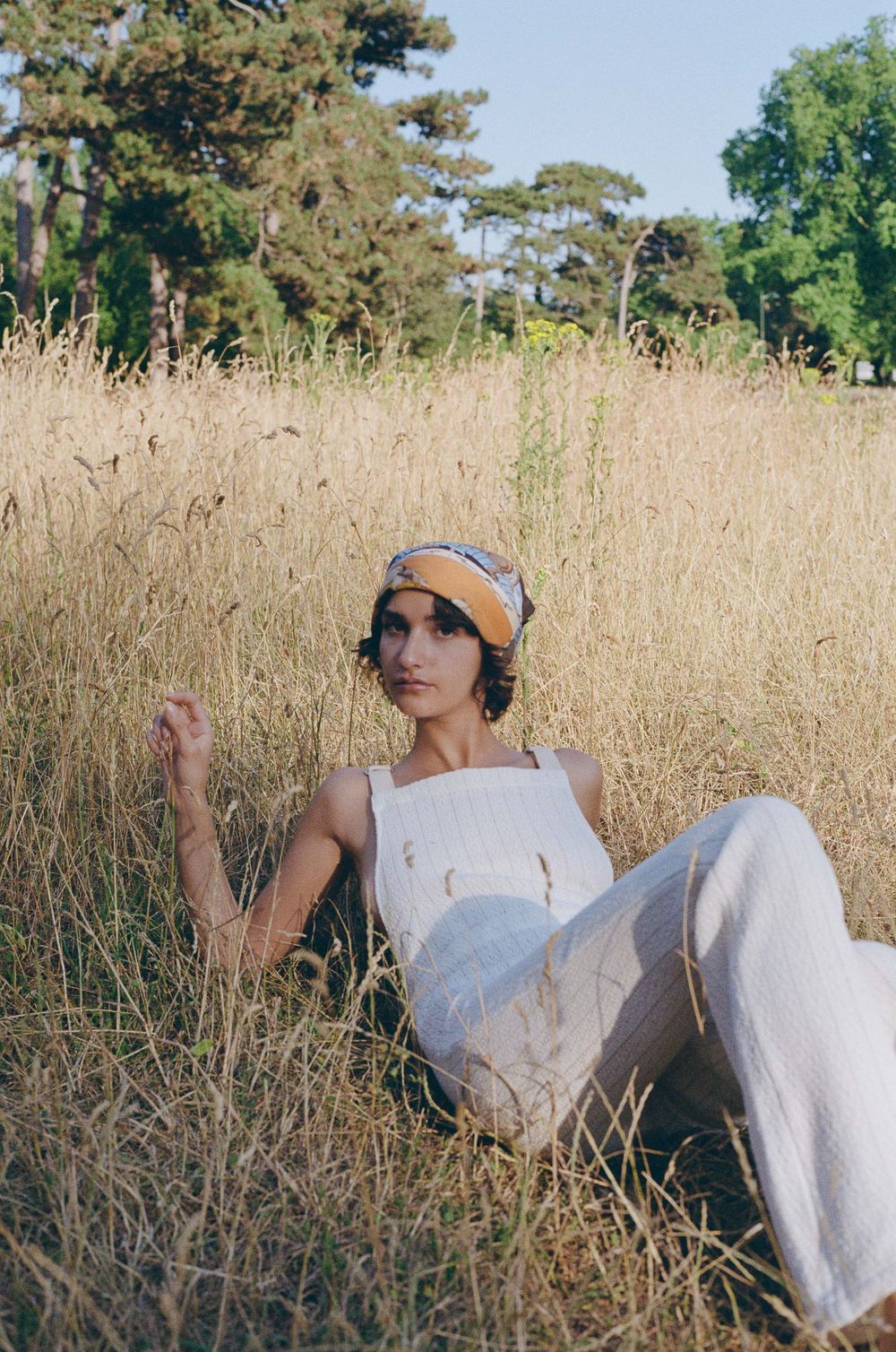 A figure in a white outfit and headband reclines in a field of tall golden grass with trees in the background.