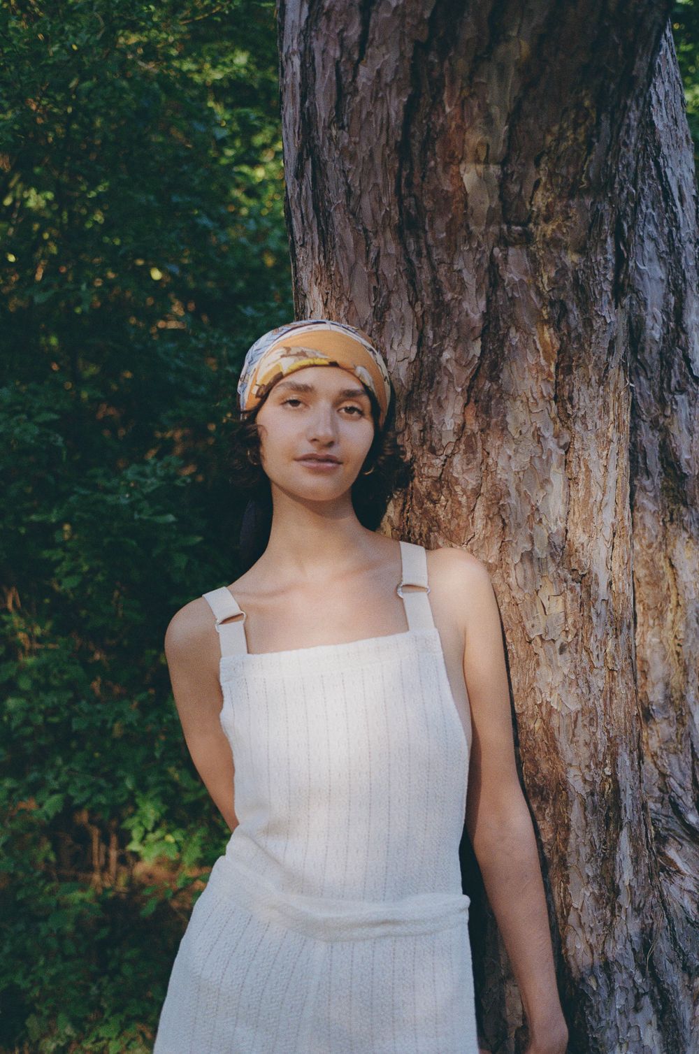 A figure in a white sleeveless dress stands next to a large tree trunk in a wooded outdoor setting.