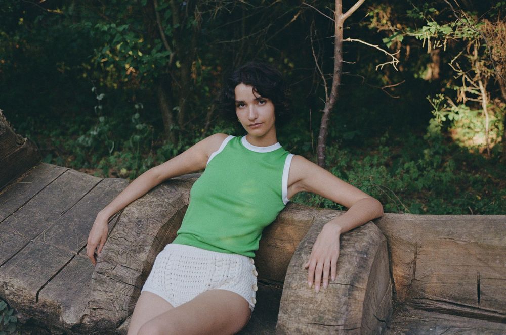 Person in green tank top and white shorts lounges on rustic wooden bench surrounded by lush forest scenery.
