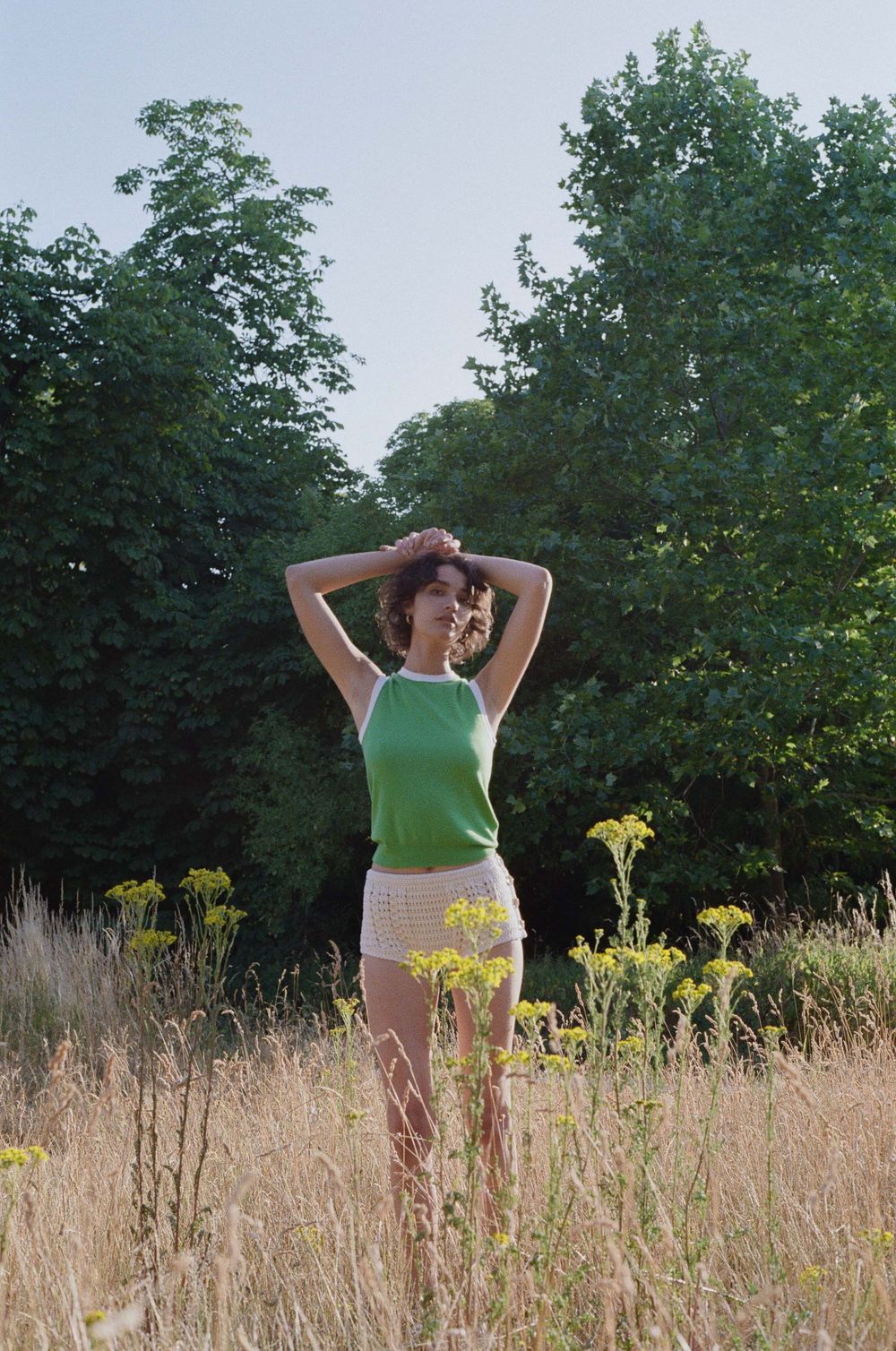 A dancer in a green top and yellow skirt performs a graceful pose in a sunny meadow filled with tall grass and trees.