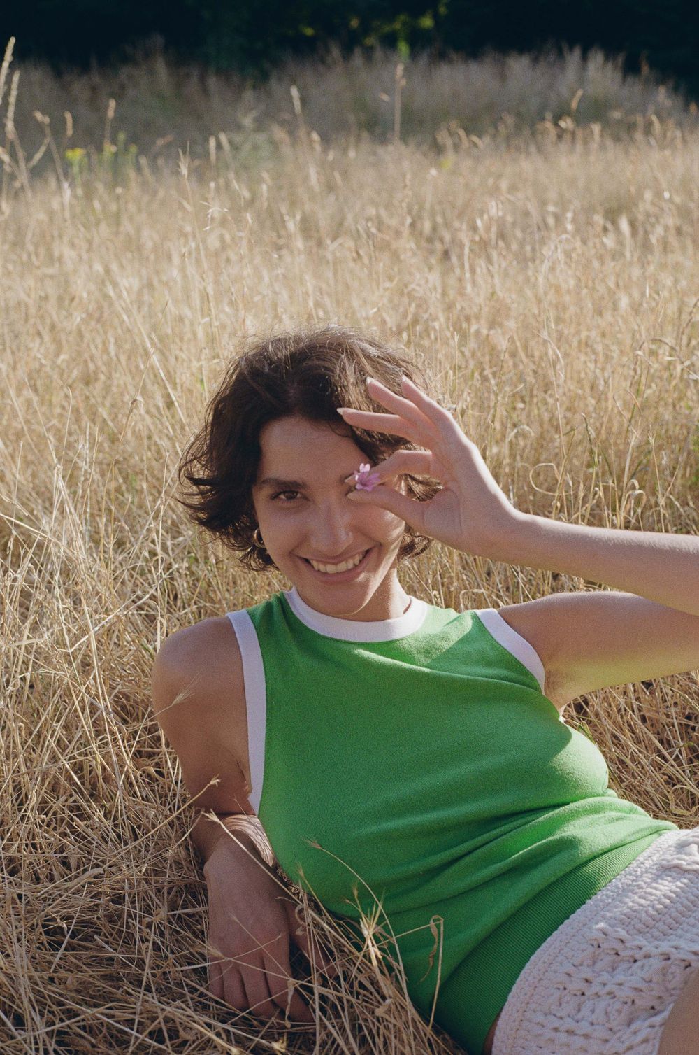 Someone wearing a green tank top lies in golden grass on a sunny day, holding up their hand with a playful expression.