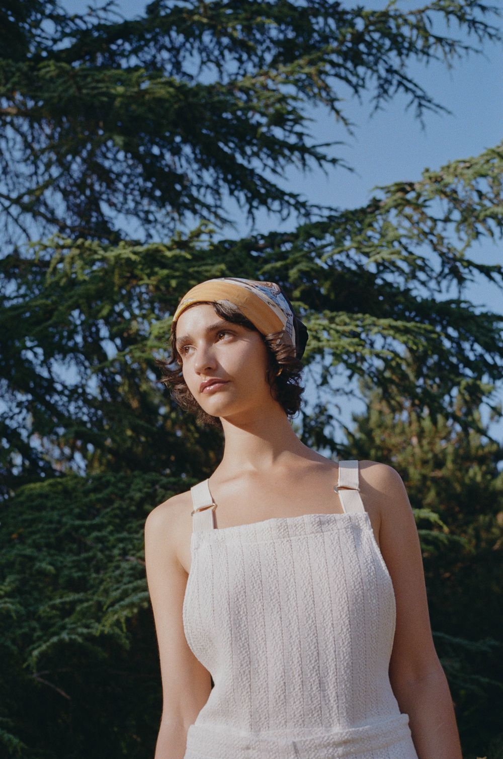 Someone in a white summer dress and beige headwrap poses outdoors against a backdrop of evergreen trees.