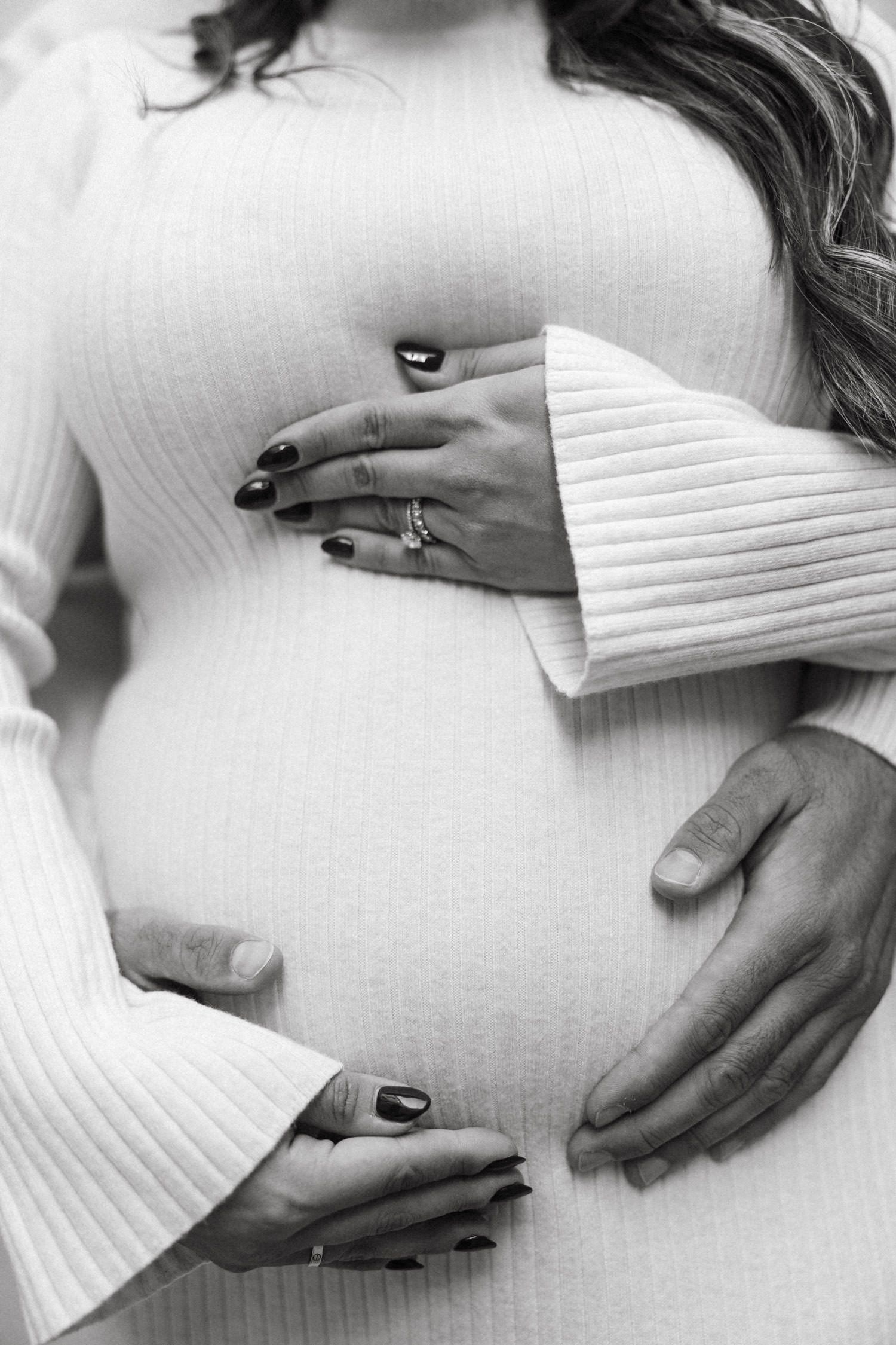Close-up of hands cradling a pregnant belly in white knit dress.