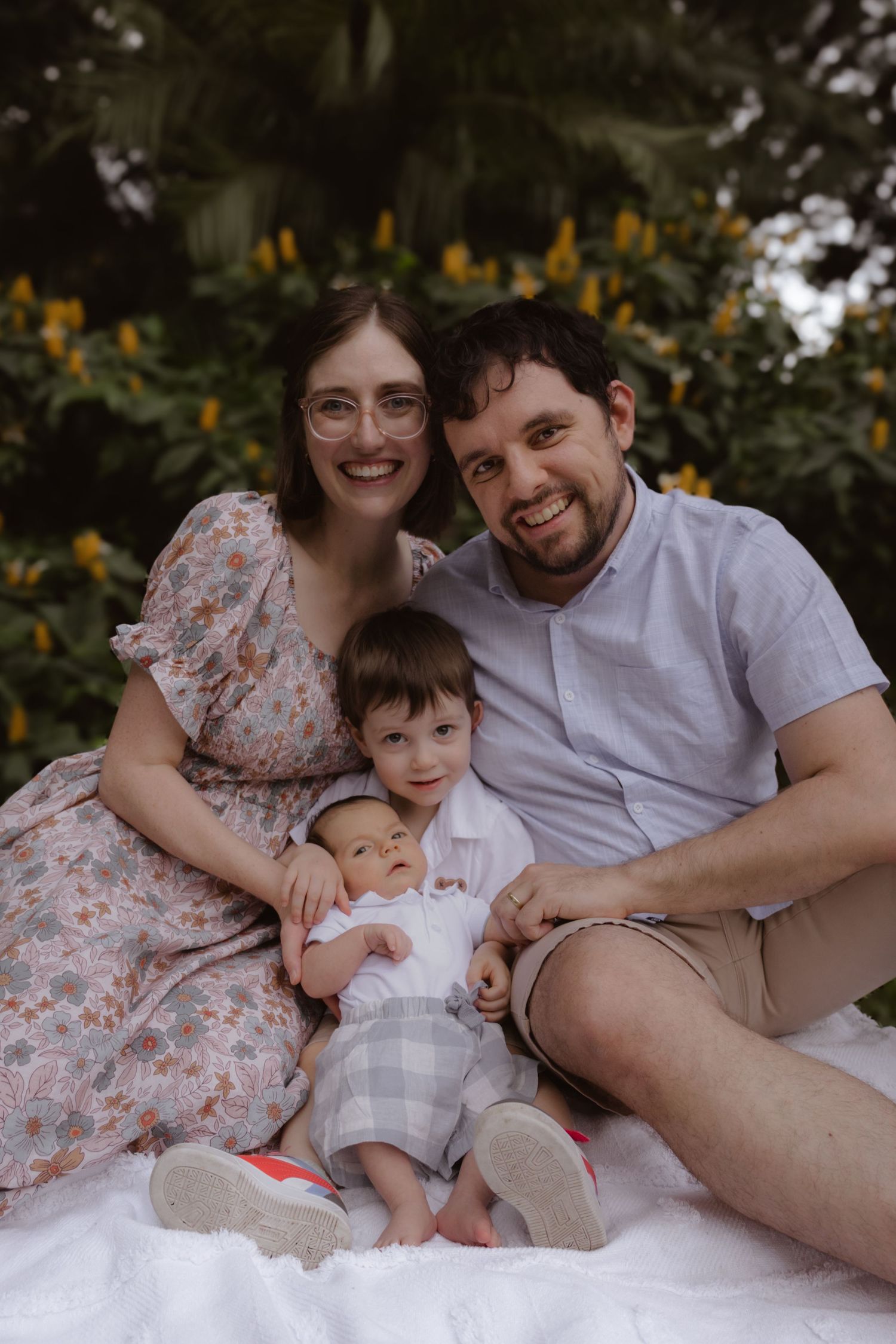 Family relaxing together on a white blanket surrounded by blooming flowers in a garden setting.