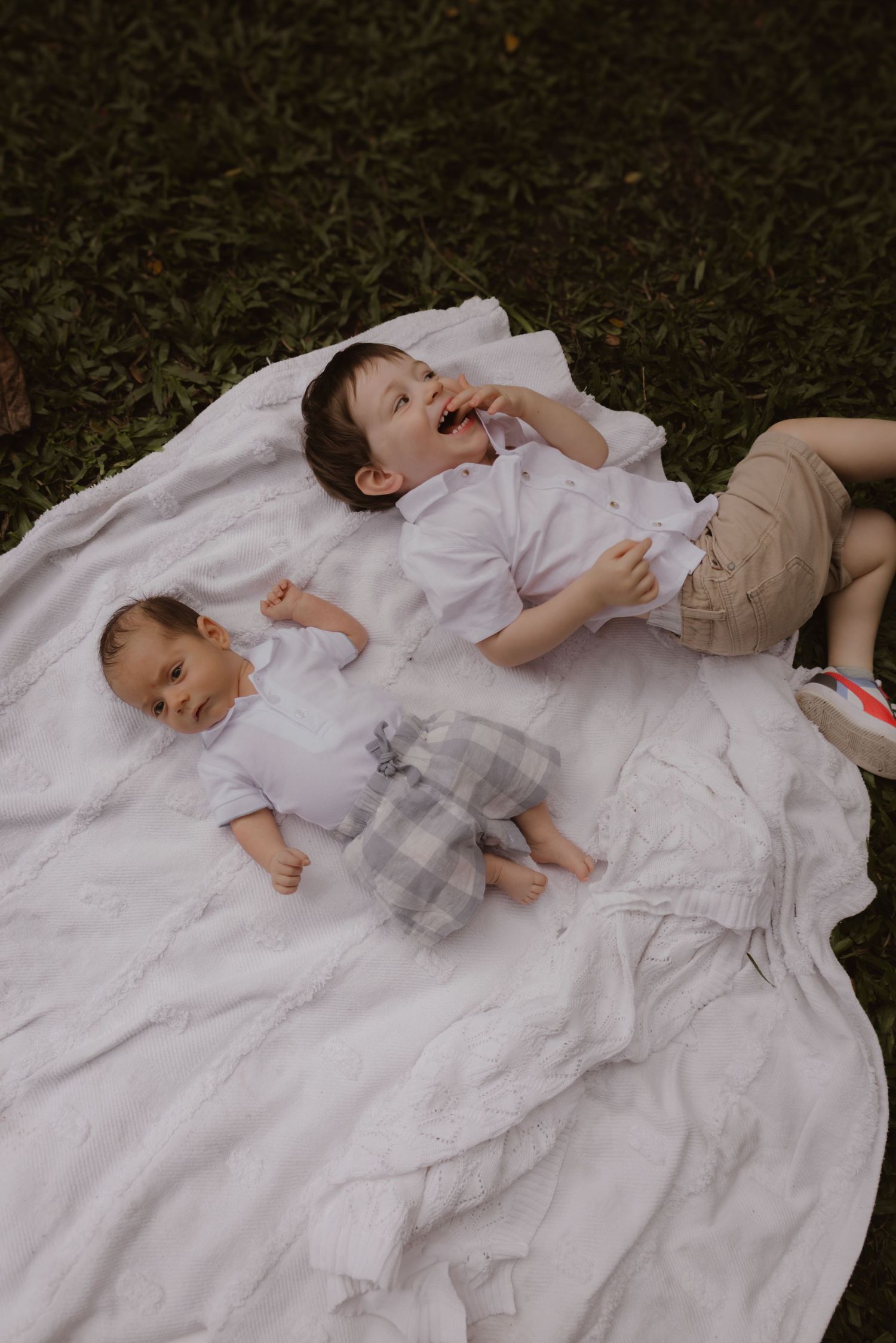Baby lying on white blanket in grass wearing delicate white dress and ruffles.