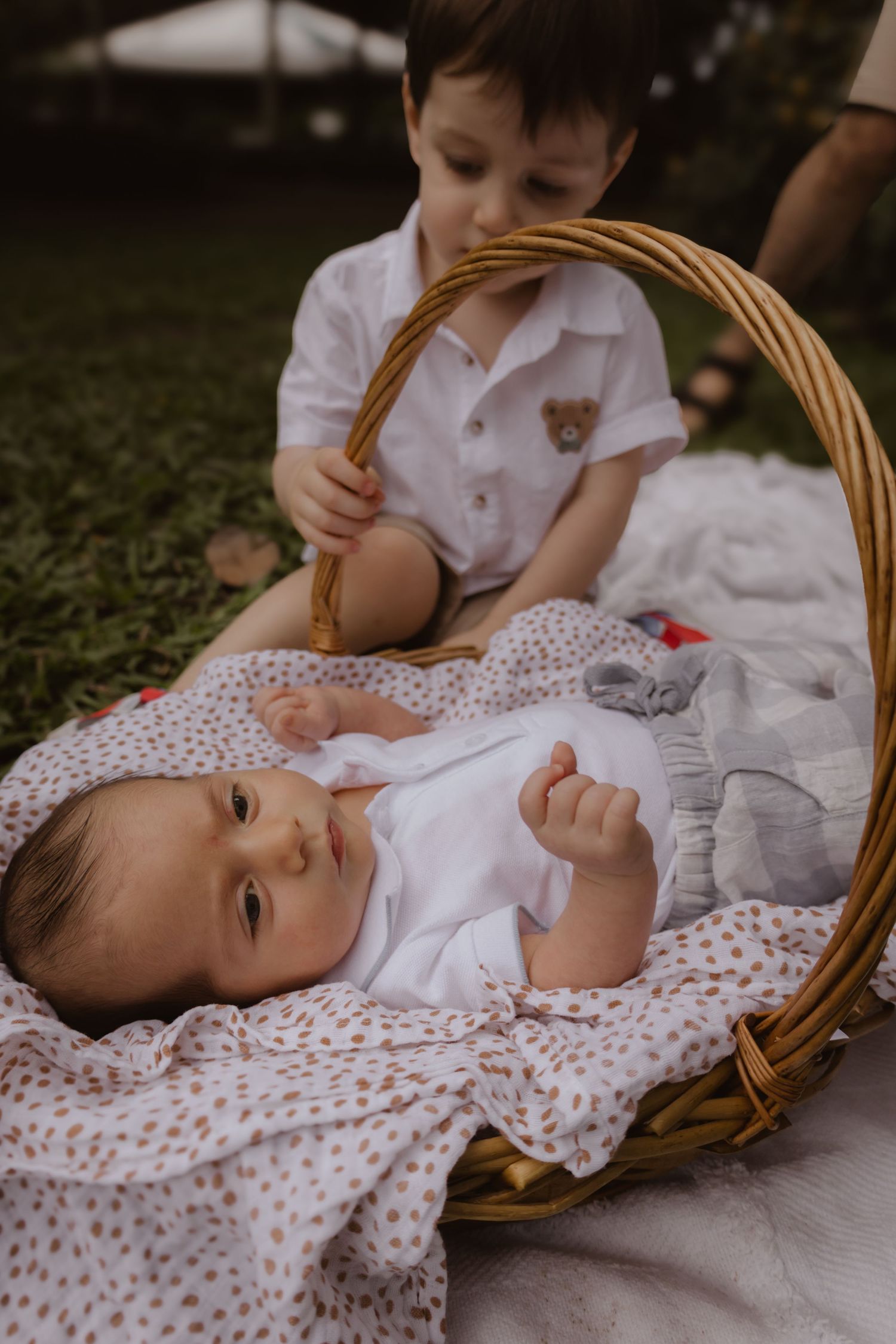 Baby resting in wicker basket on white blanket wearing polka dot outfit.
