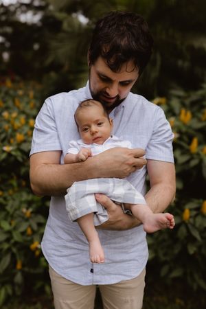 Parent in white shirt holding infant outdoors with sunflowers in background.