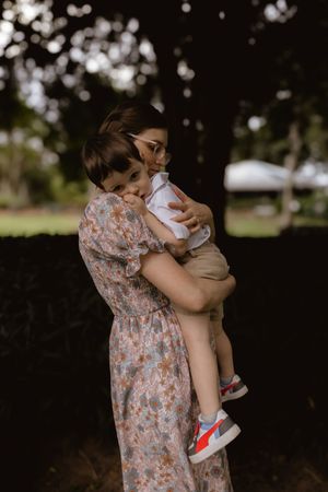 Parent in floral dress embracing baby outdoors under trees.