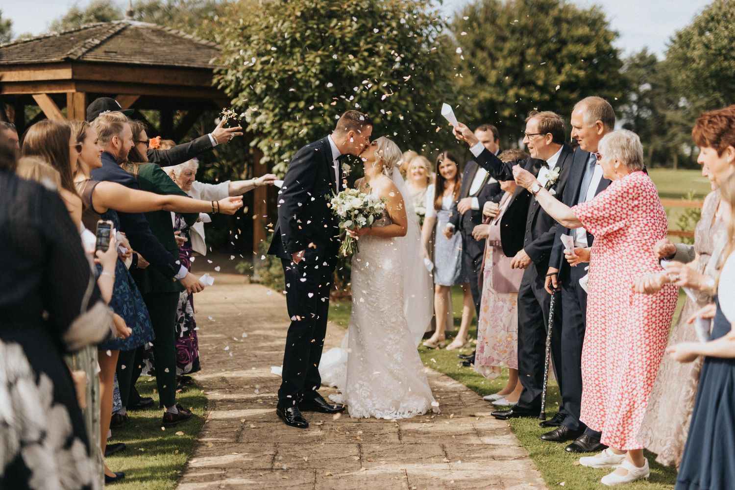Wedding guests throw confetti over the couple during an outdoor garden ceremony on a stone pathway.